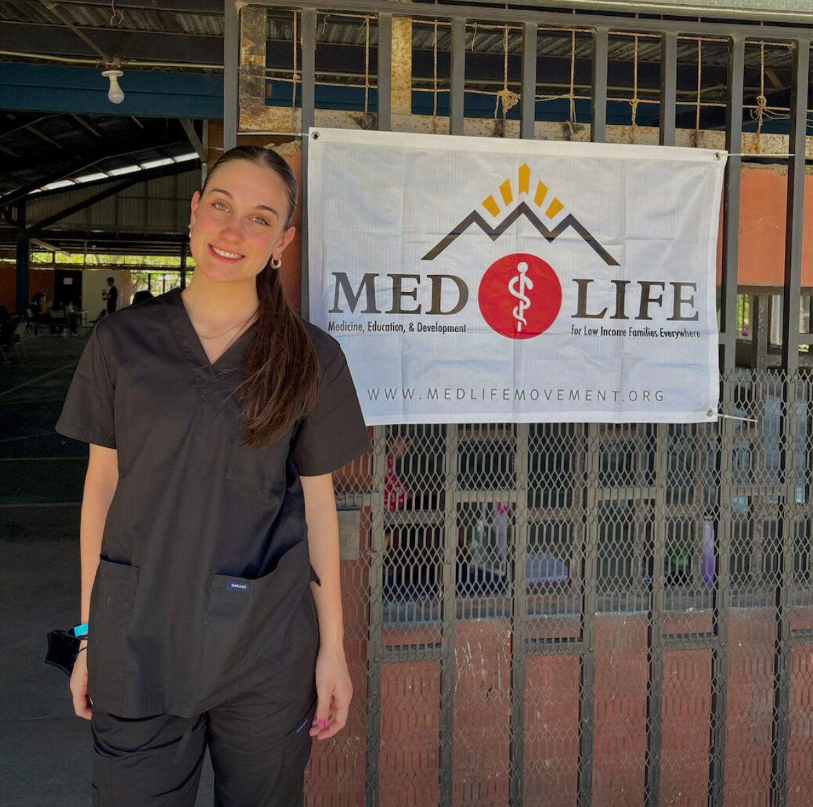 photo of Emily DiCesare, standing in front of a medical clinic in Costa Rica. She is wearing scrubs and behind her is a poster saying "MEDLIFE", promoting good healthcare for low-income families